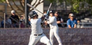 Baseball batter in white uniform swings at a pitch on a dirt infield with spectators in the background.