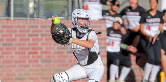 Softball catcher in white pinstriped gear and helmet running on the field, with a brick wall and cheering spectators in the background.