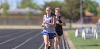 Female runner in gray and blue sprinting on outdoor track, leading a group of competitors behind her.