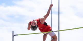 Male pole vaulter in a red uniform clears a bright green bar outdoors against a blue sky.