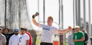 Male athlete in a white shirt (Bushland) releasing a shot put during a field event, with spectators behind a safety net.