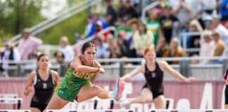 Female athlete in a green uniform clearing a hurdle mid-race at a track meet, with blurred crowd in the stands behind her.