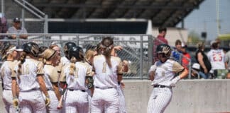 Softball players in white pinstripe uniforms huddle on the field before a game.