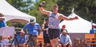 Male shot-put athlete mid-throw during a track meet, wearing a white patterned singlet and red sneakers, with officials and tents in the background.