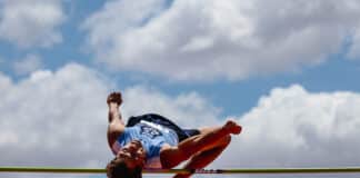 Athlete in a blue uniform clears a track hurdle midair against a blue sky with clouds.