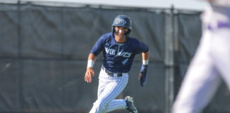 Baseball player in blue uniform running on the field, helmet on, with a blurred figure in foreground on the right.