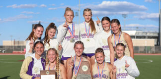 Group of female track athletes posing on a field, wearing matching uniforms and medals after a win, holding trophies.