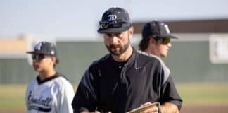 Baseball coach in a black uniform holds a clipboard and notes while two players practice in the background on a sunny field.