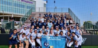 Girls softball team poses on the field holding a 'Regional Semifinal Champions' banner with a trophy in front of bleachers and fans in blue.