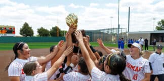 Girls softball team lifts a gold trophy in celebration on the field, wearing white uniforms with red pinstripes.