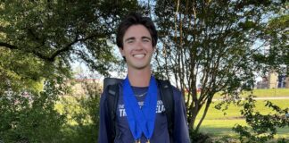 Smiling young man outdoors wearing a blue ribbon medal necklace and a backpack, with trees in the background.