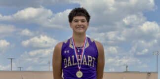 Male student-athlete in a purple Dalhart jersey with a medal around his neck, smiling on a track field against a blue sky with clouds.