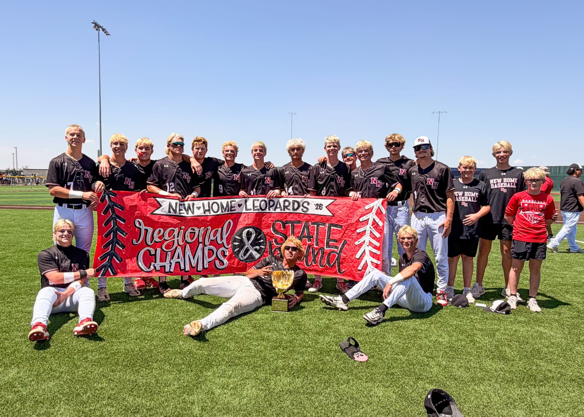 Group of young baseball players in black uniforms pose behind a red banner on a sunny field, trophy in front, celebrating a win.