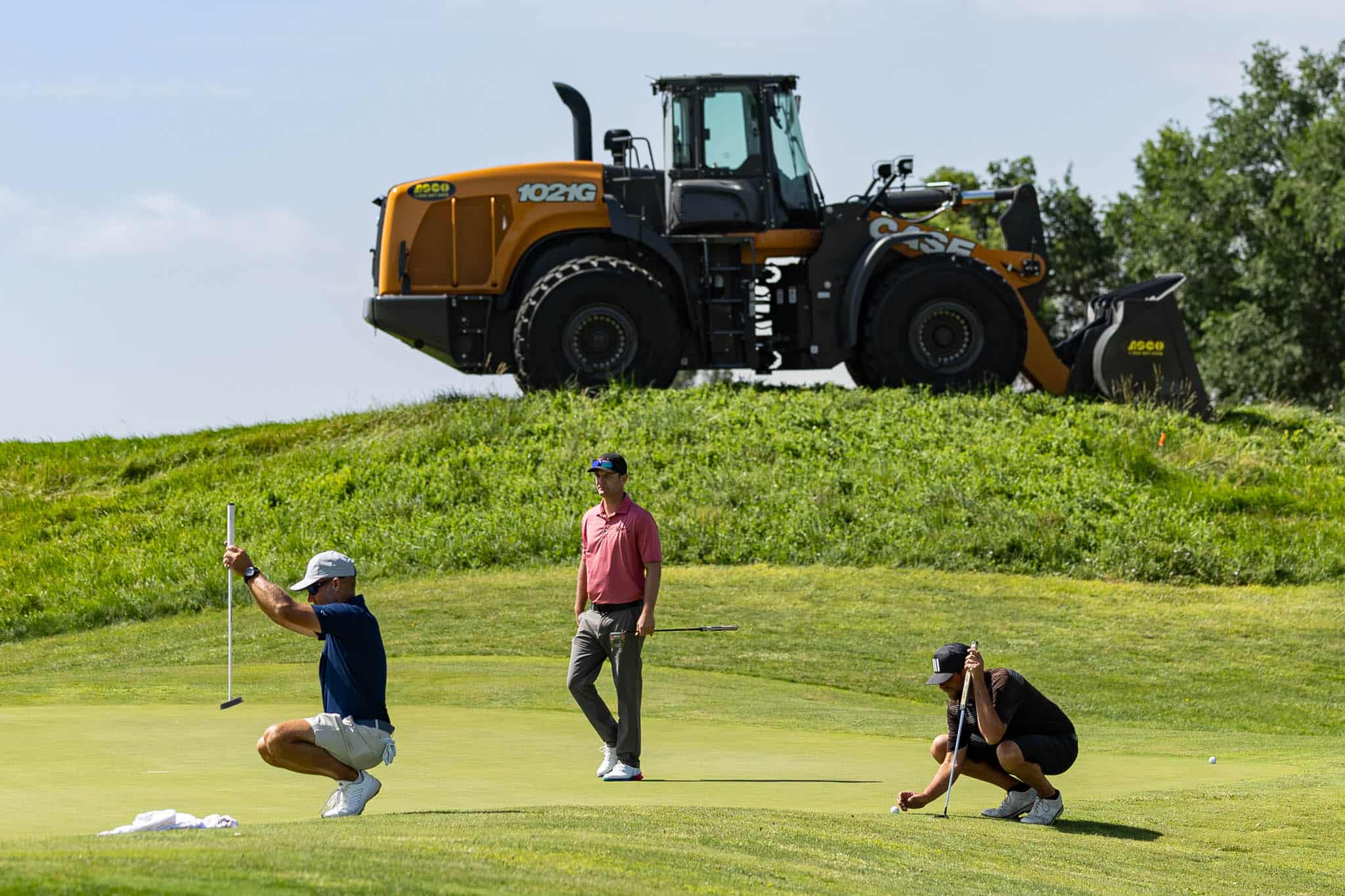 ASCOABC7-Golf-42 | Press Pass Sports Golfers on a green putting while a large orange excavator sits on a hill behind them.
