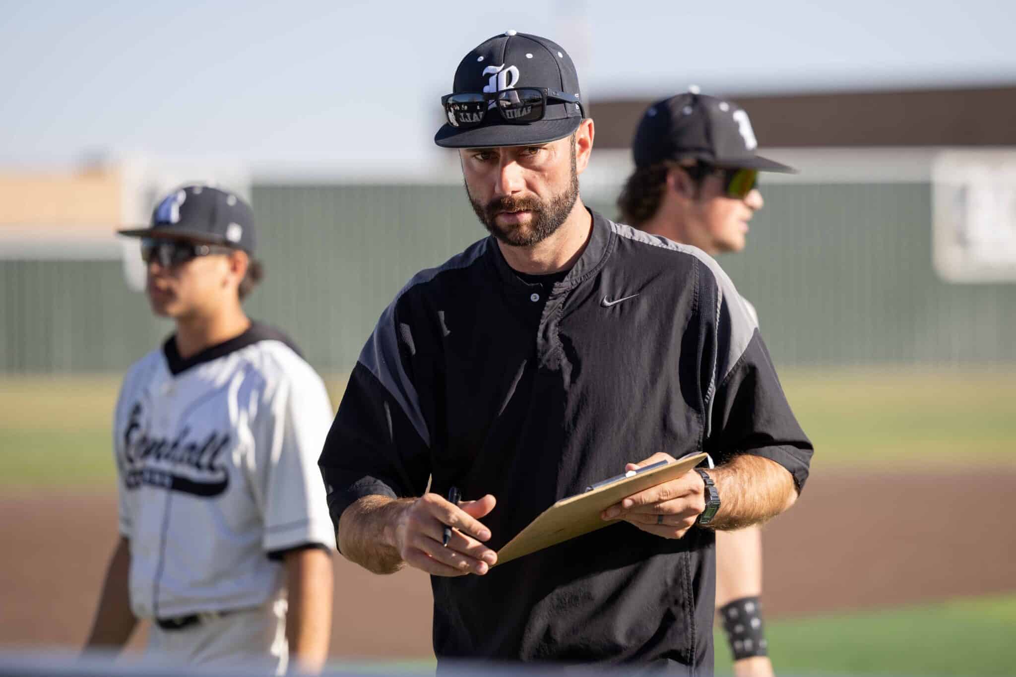 CHS-RHS-BB-8 1 | Press Pass Sports Bearded baseball coach in a black jacket holding a clipboard, with two players blurred in the background on a sunny field.