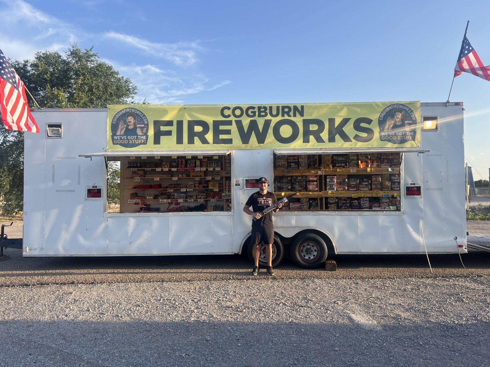 IMG_6329 | Press Pass Sports Fireworks trailer with a bright yellow banner reading'COGBURN FIREWORKS' and American flags on both ends; a person in front holding a rocket-style firework.