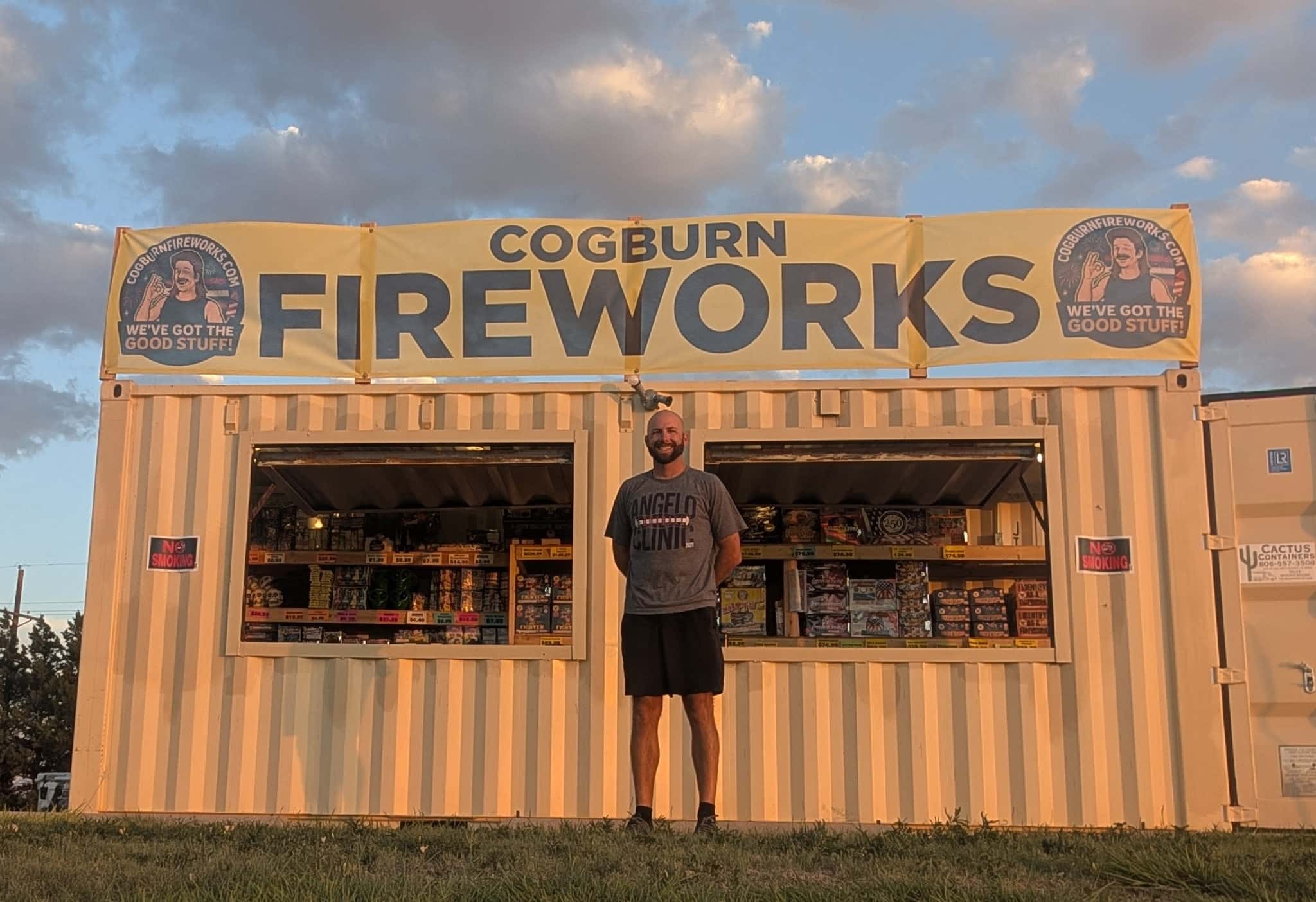 Man stands on a platform in front of a beige shipping-container storefront under a large yellow banner reading'COGBURN FIREWORKS' with circular logos on both ends.