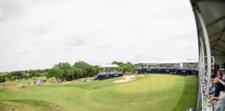 Wide-angle view of a golf tournament with spectators in tiered stands along the right side and a grassy fairway below.