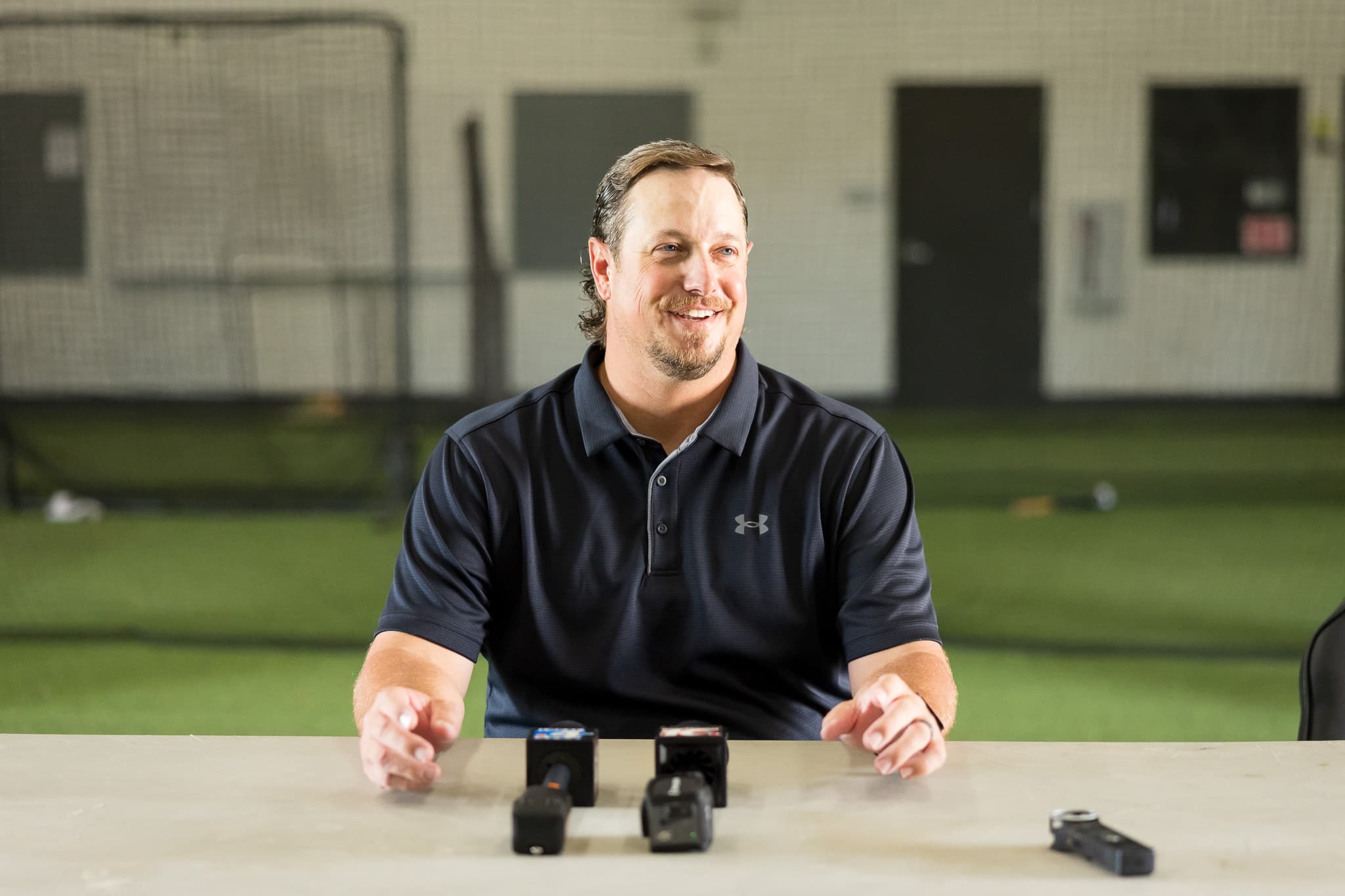 Man in a dark polo sits at a table with several small microphones in front, smiling, indoors at a sports facility.