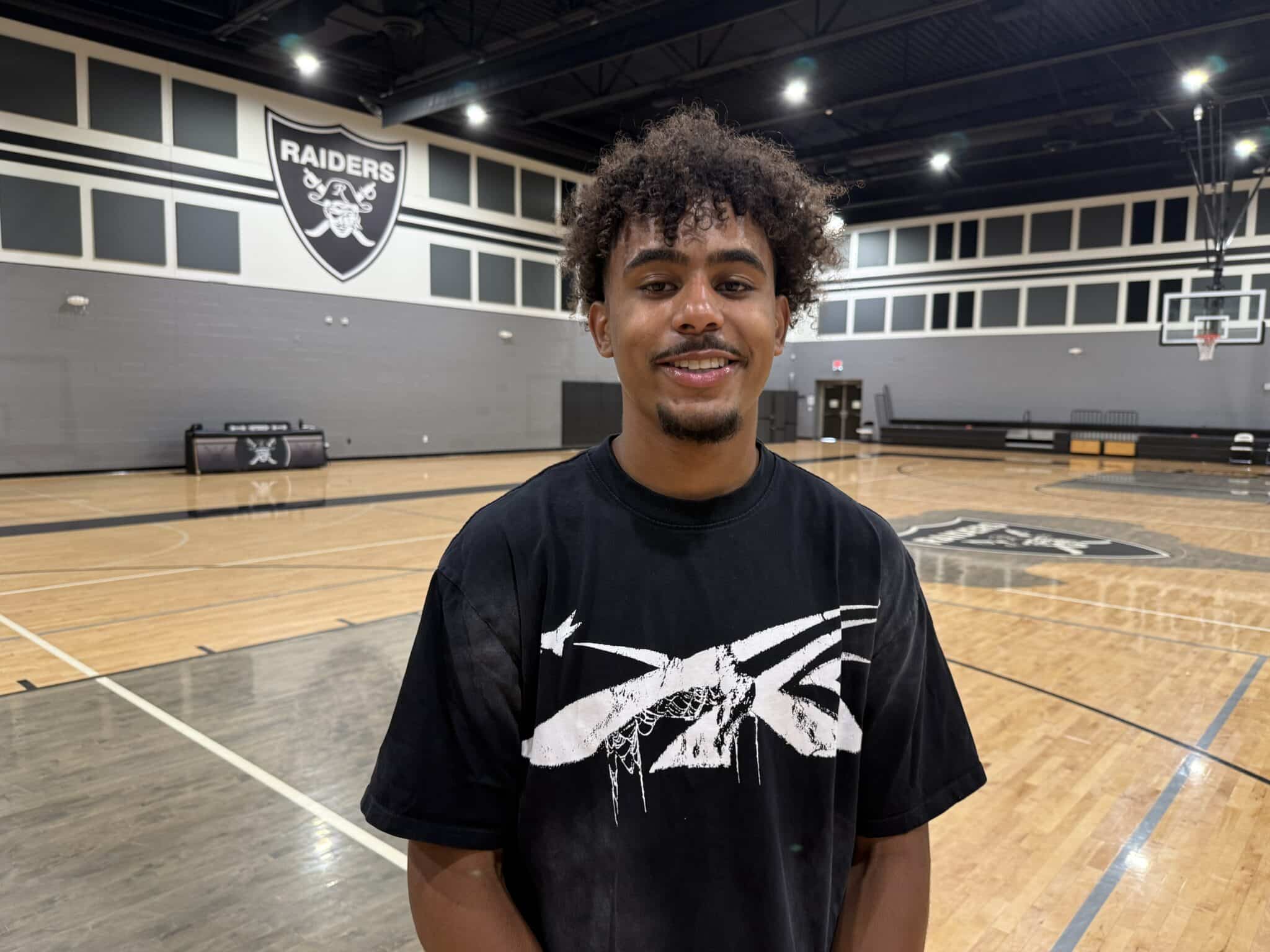 Smiling young man with curly hair in a black graphic T-shirt stands in a basketball gym with a Raiders emblem on the wall behind him.