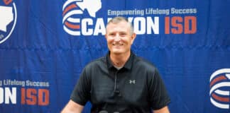 Smiling man in a black polo speaks at a press conference, microphones on the podium, blue banner backdrop with logos behind him.