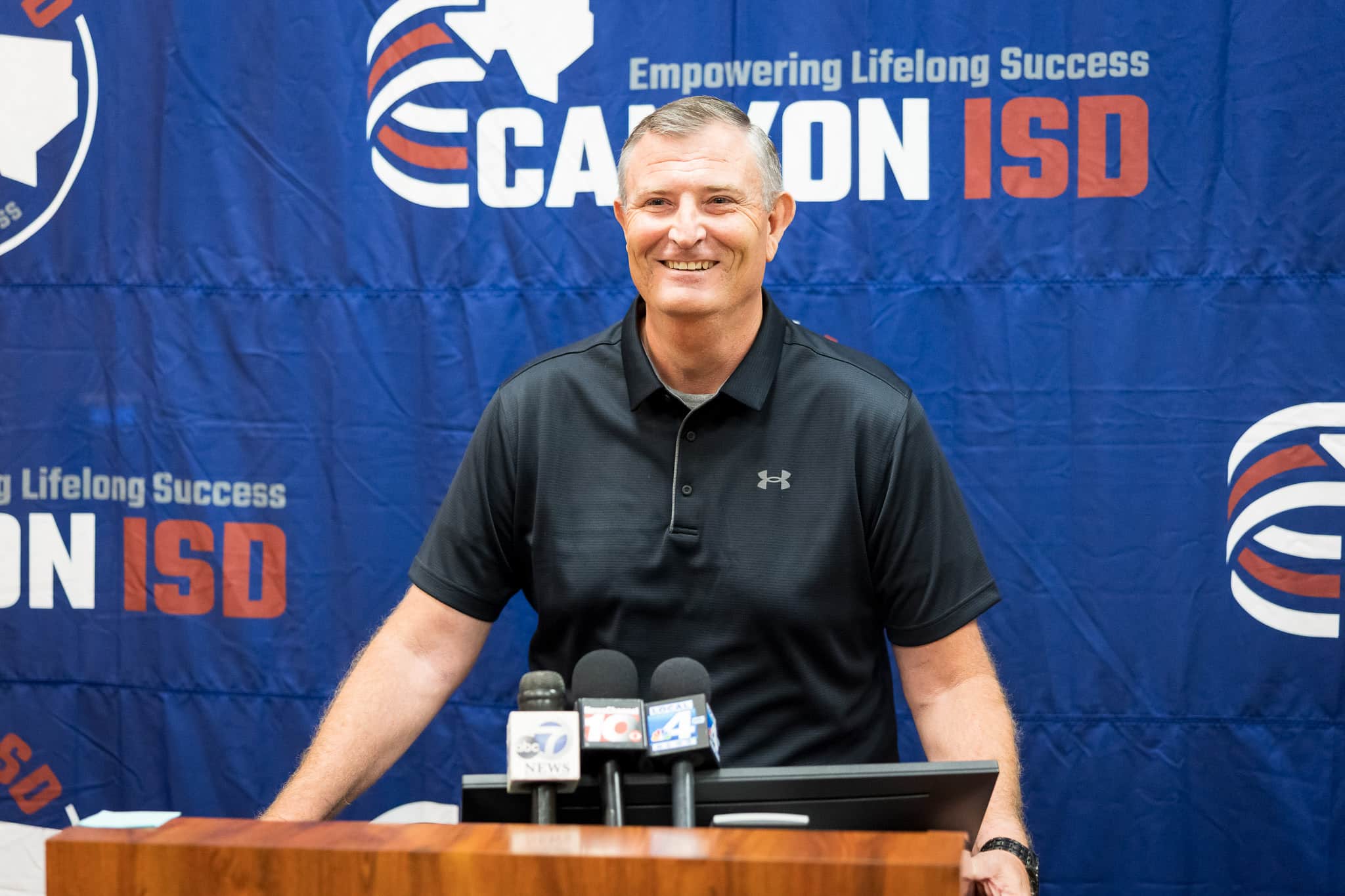 Smiling man in a black polo speaks at a press conference, microphones on the podium, blue banner backdrop with logos behind him.