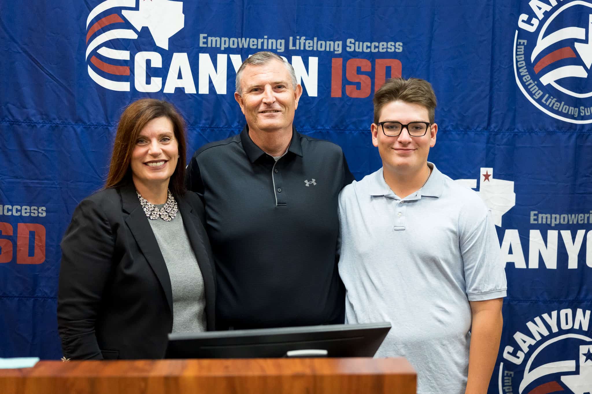 Three people—woman on left, man in middle, and younger man on right—smile in front of a blue backdrop that reads'Empowering Lifelong Success' with large white and red lettering.